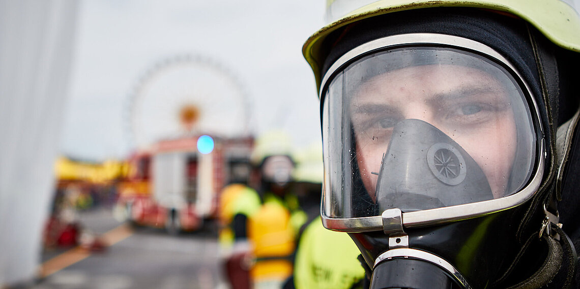 Nahaufnahme eines Feuerwehrmanns mit Atemschutzmaske und Helm, unscharfer Einsatzhintergrund mit Feuerwehrfahrzeug
